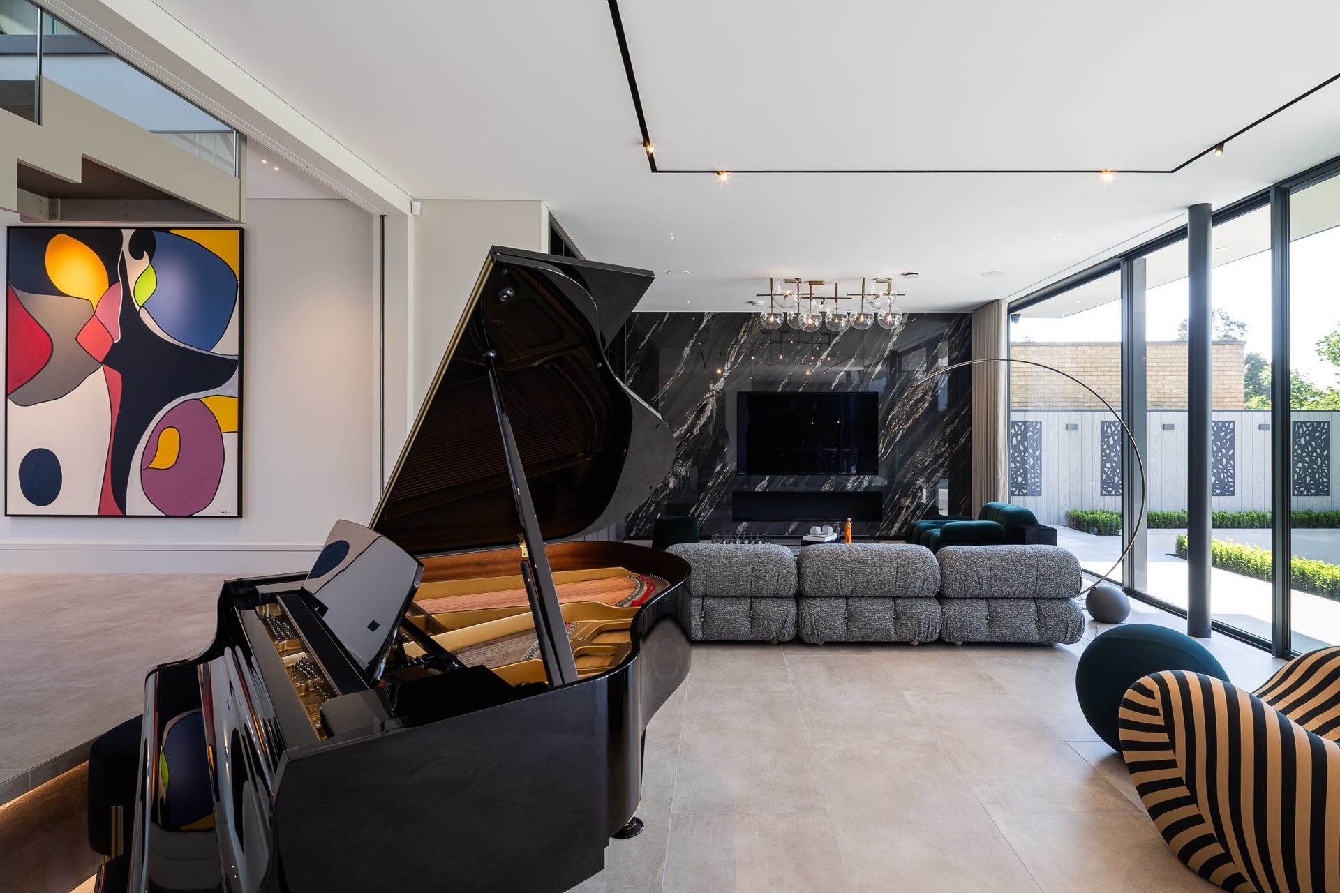 Open plan living room with grand piano as focal point, bold abstract artwork, marble feature wall with integrated TV, grey modular sofa and floor-to-ceiling glazing